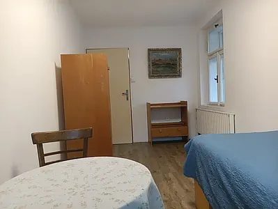 Zařízený pokoj s dřevěným nábytkem, stolkem v popředí, postelí s modrým přehozem a oknem